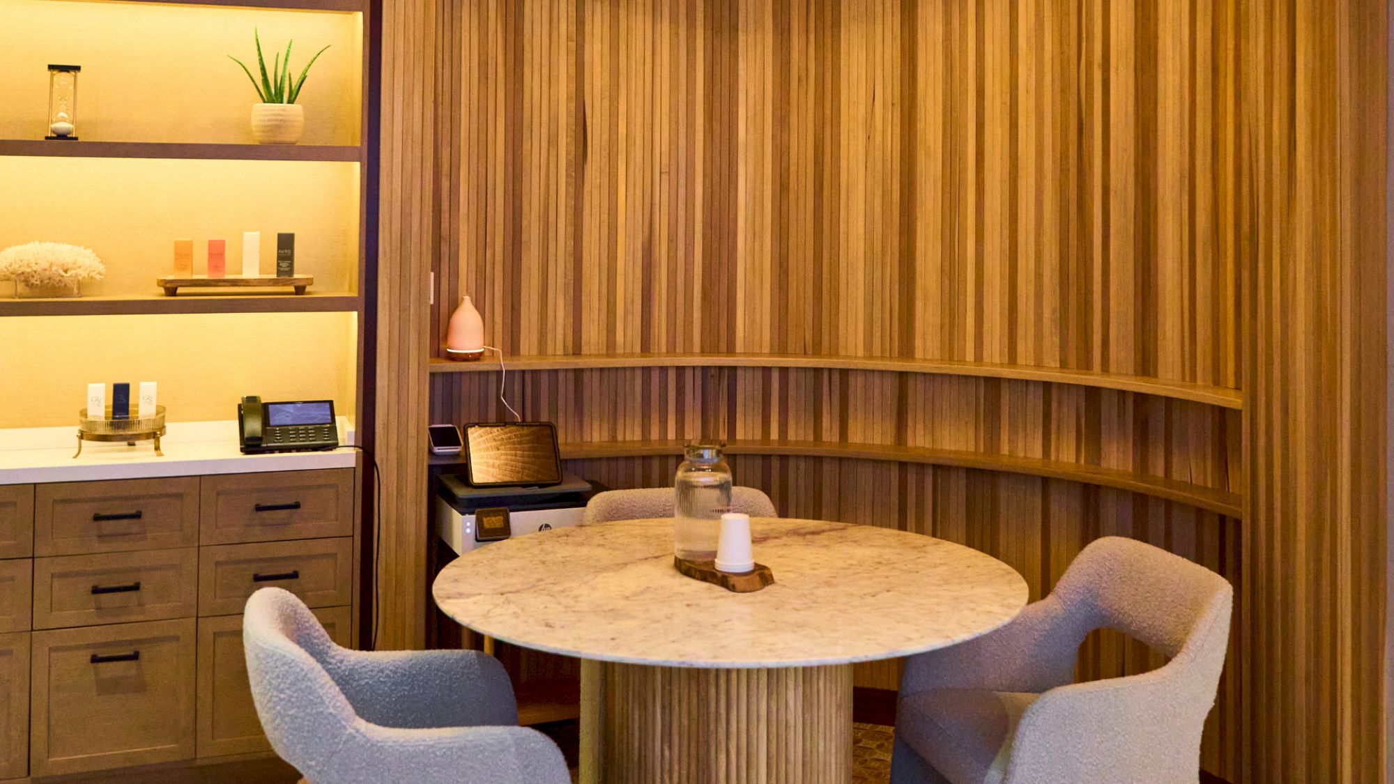 A cozy, wood-wrapped seating nook with a round marble table, two soft chairs, built-in shelving, and warm, ambient lighting.