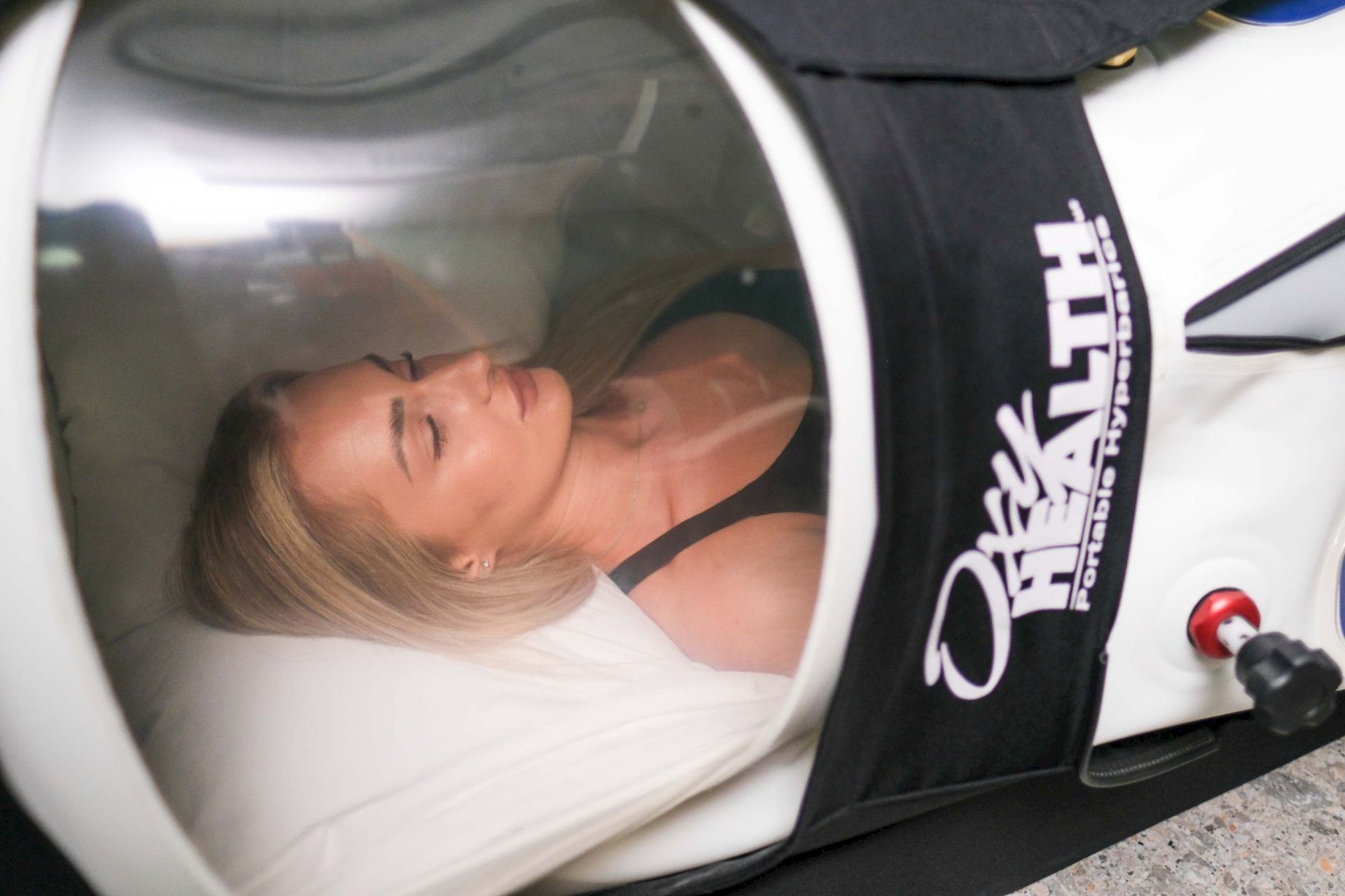 A blonde woman reclines inside a white machine or seat, with a black panel beside her featuring printed text and icons.