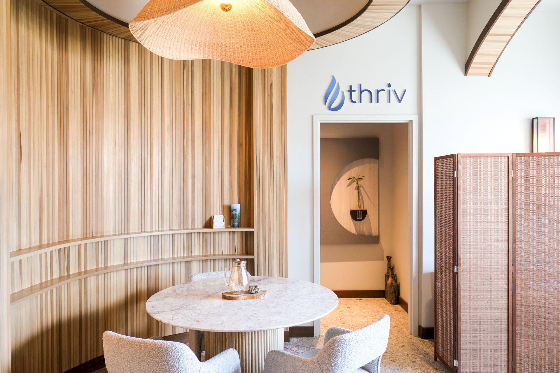 Bright office lounge with wooden slats, a marble table, pendant lamps, and the &ldquo;thriv&rdquo; logo on the wall.