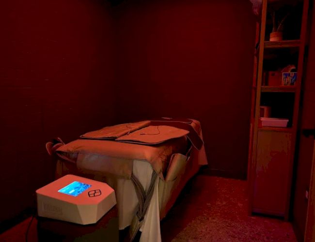 A dimly lit room with a single bed, a turntable-like device on a nearby table, and a tall cabinet; the bed is neatly made with a blanket.