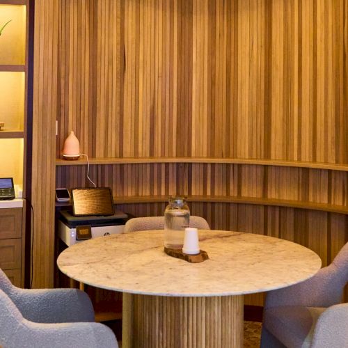 A cozy spa lounge with a round marble table, plush chairs, and wooden wall shelves holding toiletries and decor.