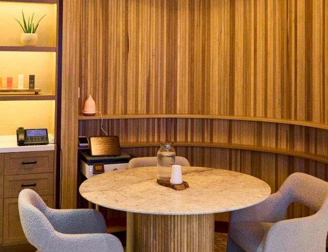 A cozy spa lounge with a round marble table, plush chairs, and wooden wall shelves holding toiletries and decor.