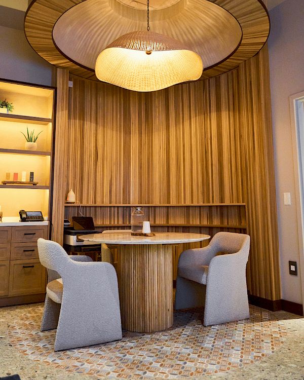A stylish circular wood dining nook with a large layered pendant lamp, striped wood panels, built-in shelves, and two gray upholstered chairs, cozy and modern.