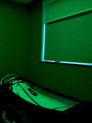 A dark room with green lighting, a single bed or couch, and a window with a green blind; eerie, moody atmosphere.