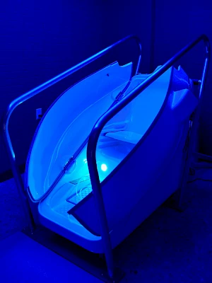 A futuristic-looking white tub or cradle on a metal stand, glowing blue under dim lighting, with rails and hoses suggesting a spa or float pod.
