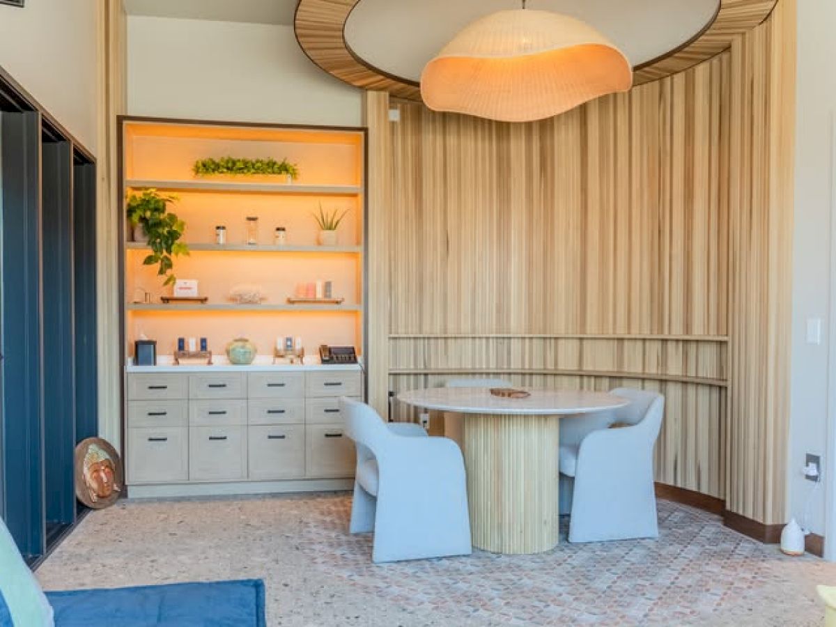 A stylish, sunlit dining nook with a round wooden table, light gray chairs, and a built-in wooden wall panel; a display cabinet with plants on shelves sits to the left, plus a warm hanging lamp overhead.