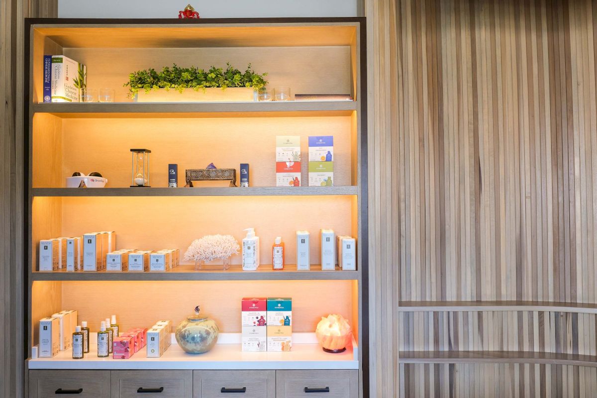 A spa display shelf filled with skincare bottles and boxes, greenery, glasses, books, and a glowing salt lamp.