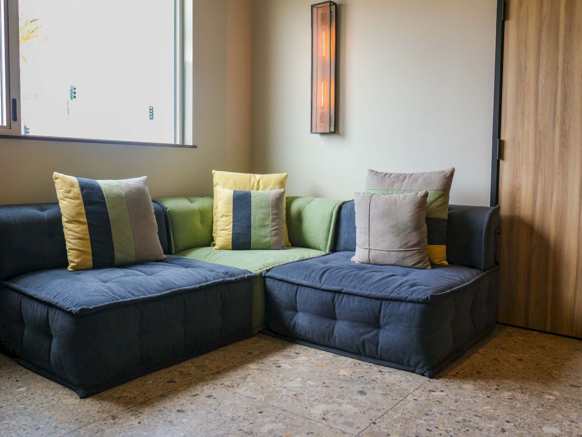 A modern living room with an L-shaped sofa, colorful pillows, a wall light heater, and a window letting in daylight.