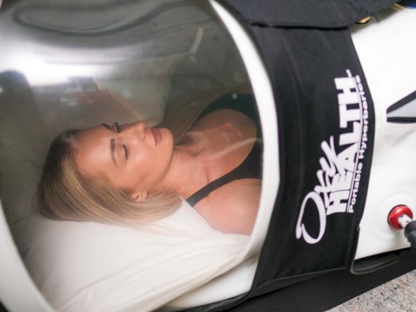 A blonde woman is reclining in a bed-like tanning or spa machine while looking at the camera in a close-up photo.