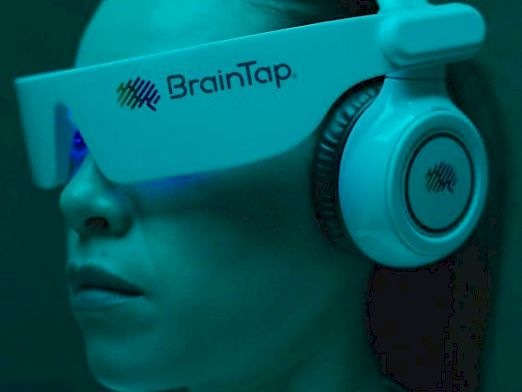 A person wearing a BrainTap-branded headband/gear in teal lighting, with headphones over the ears.